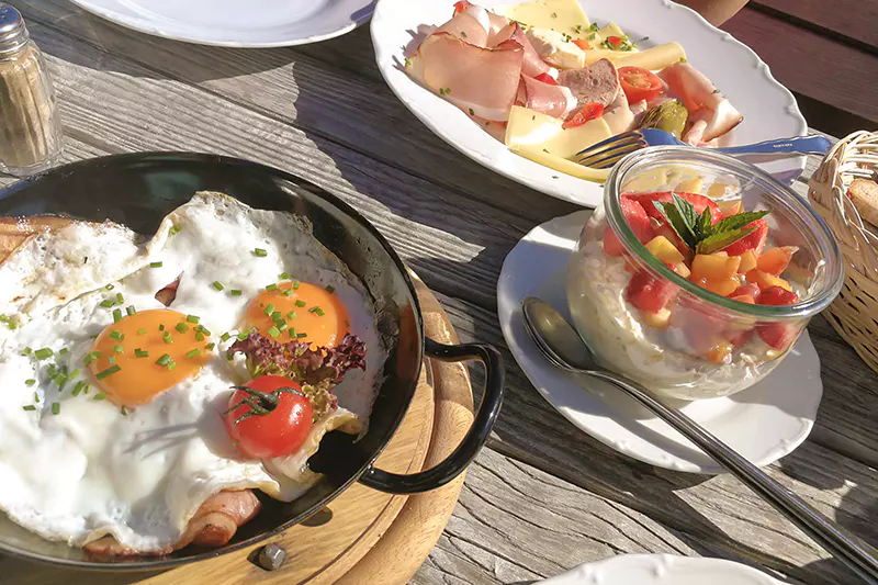 Een ontbijtbuffet met spiegeleieren, tomaten, vleeswaren met kaas en een potje yoghurt met vers fruit, geserveerd op een houten tafel in het felle zonlicht.